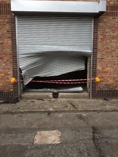 Severely buckled metal roller shutter across a brick doorway, cordoned off with red and white tape