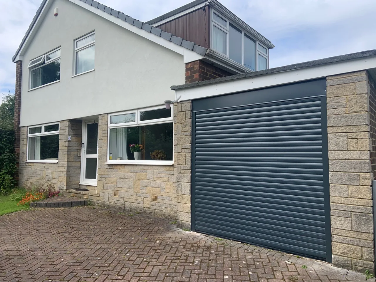 Grey roller garage door with horizontal cladding above, beside a white house.