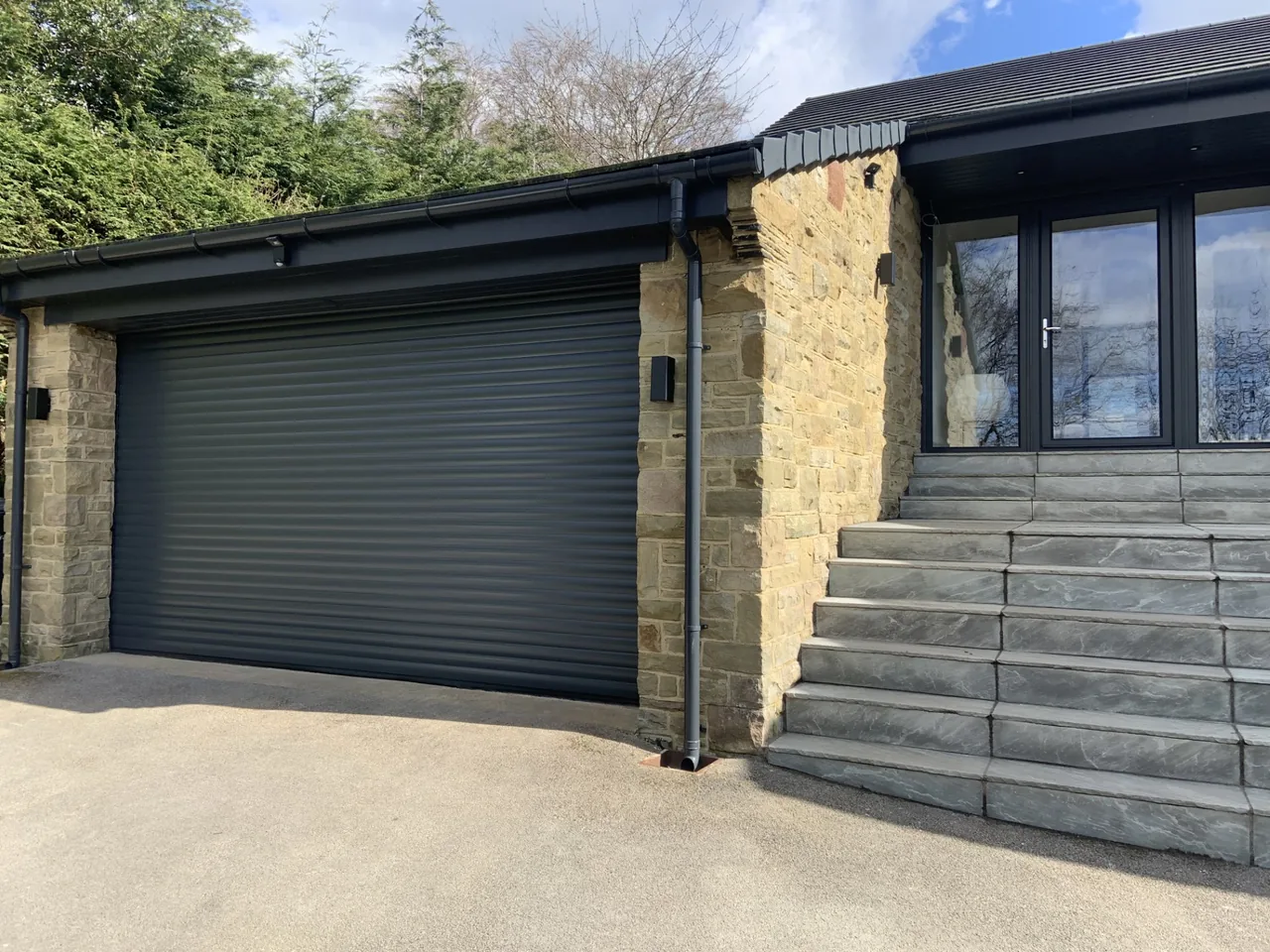 Grey roller garage door below a matching fascia beside a stone house.