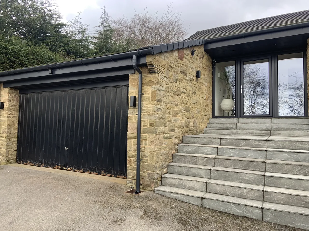 Dark garage door with vertical panels beside a stone house, before replacement.