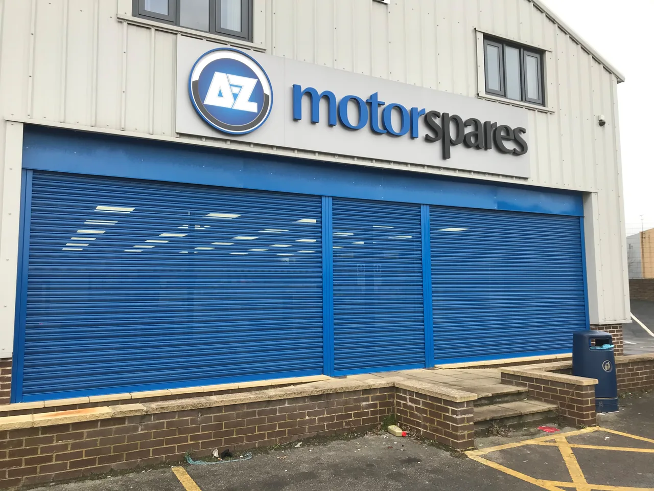 Blue roller shutters with vision panels across a motor spares shopfront