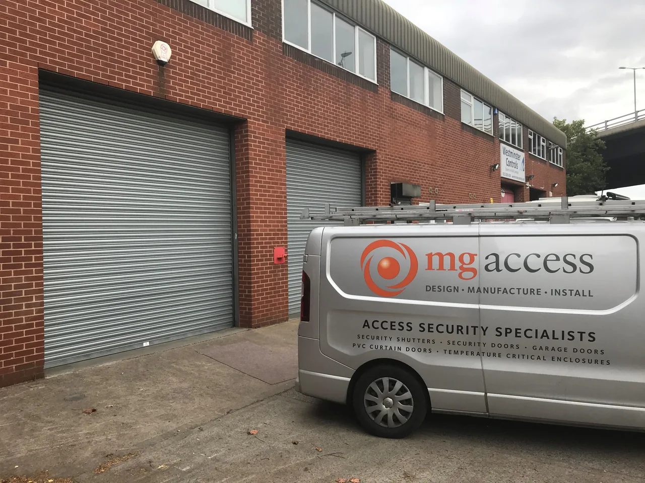 Grey roller shutter fitted to a brick industrial unit beside an MG Access van