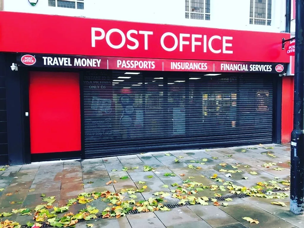 Dark perforated security shutter across a red Post Office shopfront