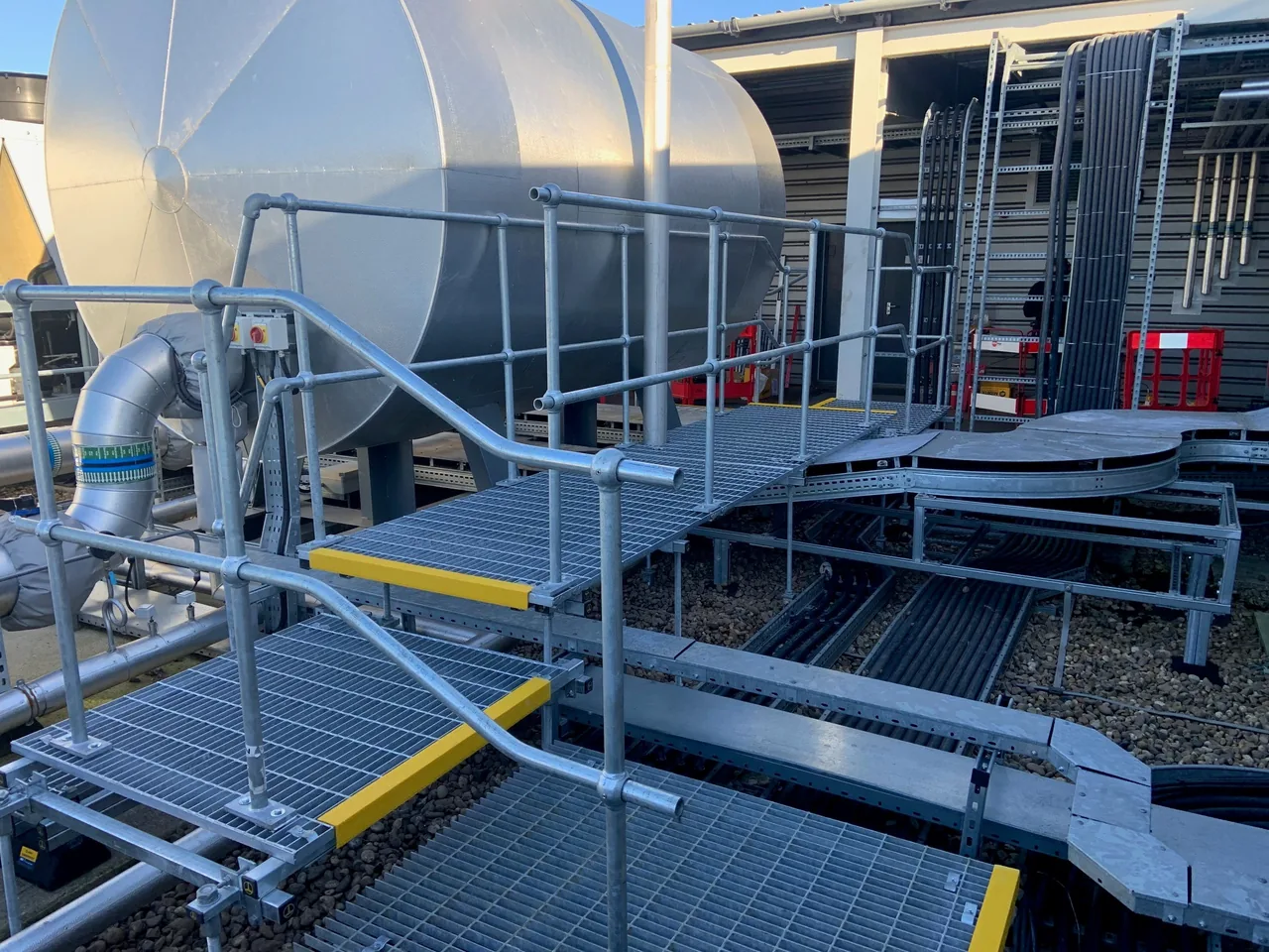 Raised metal walkway with handrails and yellow step edges beside rooftop plant