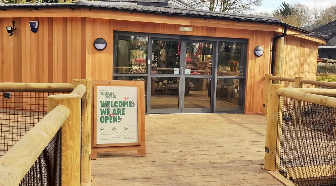 Glazed automatic entrance doors in a timber-clad shopfront, approached by a decked walkway.